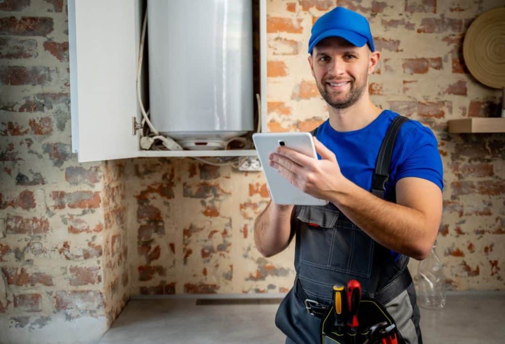 A water heater technician in protective workwear uses a tablet while inspecting a residential water heater in a utility room.