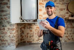 A water heater technician in protective workwear uses a tablet while inspecting a residential water heater in a utility room.